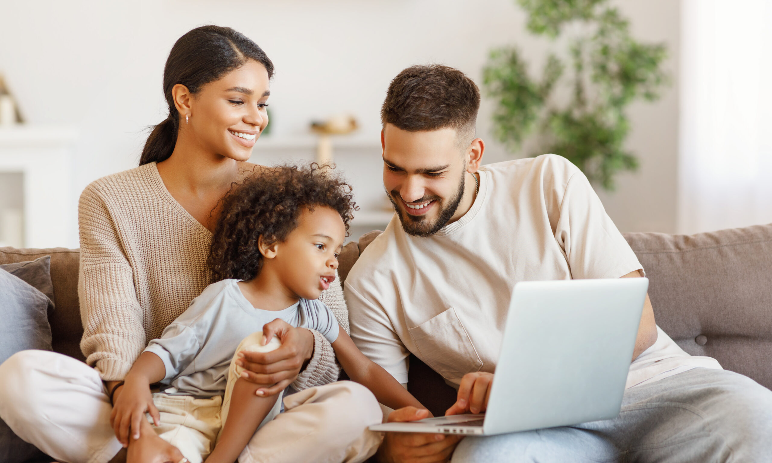 A woman, a man, and a young child sit together on a couch, smiling and looking at a laptop screen. The room is bright and cozy with plants in the background.