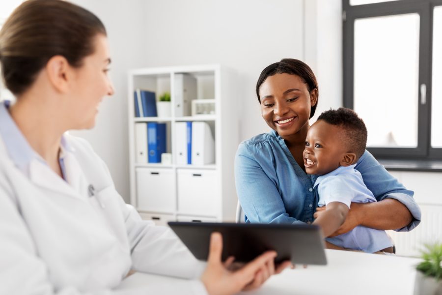 A doctor holds a tablet while talking to a smiling woman and her young son, who is sitting on her lap, in a bright medical office.