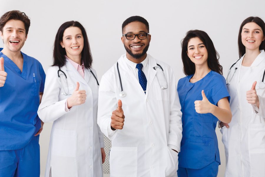 A group of five smiling healthcare professionals, including doctors and nurses specializing in family medicine in Erie PA, stand together giving thumbs up, dressed in white coats and blue scrubs against a plain background.