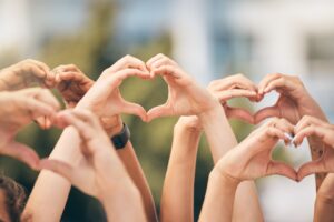 A group of people raise their hands together, each forming a heart shape with their fingers against a blurred outdoor background, symbolizing unity and support for heart health tips.