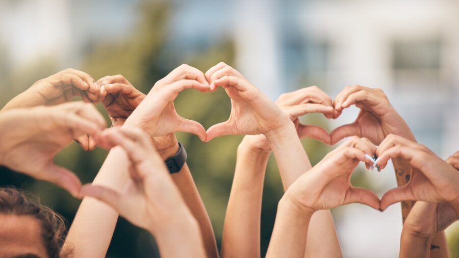 A group of people raise their hands together, each forming a heart shape with their fingers against a blurred outdoor background, symbolizing unity and support for heart health tips.