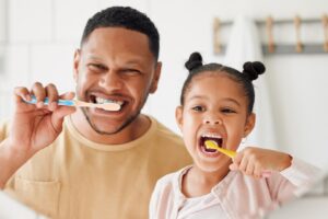 A man and a young girl smile and brush their teeth together in front of a bathroom mirror, holding toothbrushes and looking happy as they practice healthy smile habits.