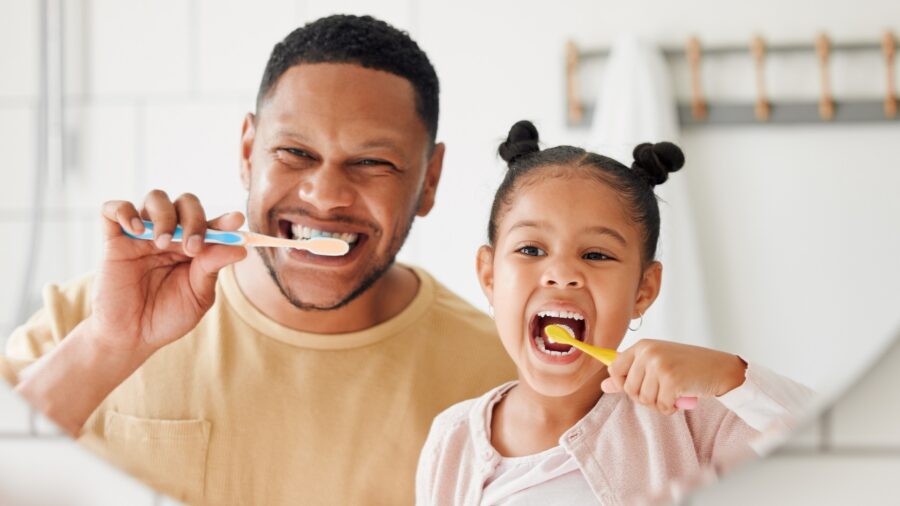 A man and a young girl smile and brush their teeth together in front of a bathroom mirror, holding toothbrushes and looking happy as they practice healthy smile habits.