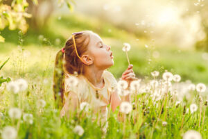 A young girl lies in a field of dandelions, blowing seeds from a dandelion puff. Sunlight filters through the grass, creating a warm, cheerful atmosphere—a gentle reminder of the importance of allergy and asthma awareness during beautiful days outdoors.