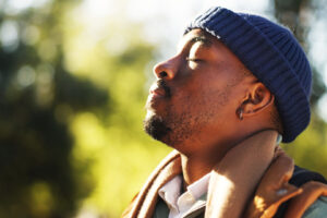 A person wearing a blue knit hat and brown jacket stands outside with eyes closed, peacefully facing the sunlight, embodying a moment of calm and mental health awareness, with a blurred green background.