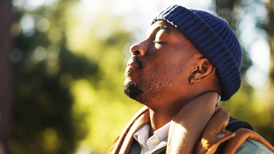A person wearing a blue knit hat and brown jacket stands outside with eyes closed, peacefully facing the sunlight, embodying a moment of calm and mental health awareness, with a blurred green background.