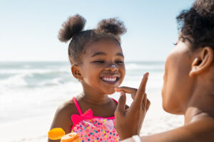 A child in a polka-dot swimsuit smiles at the beach as an adult applies sunscreen to her nose, showcasing one of the important summer safety tips.