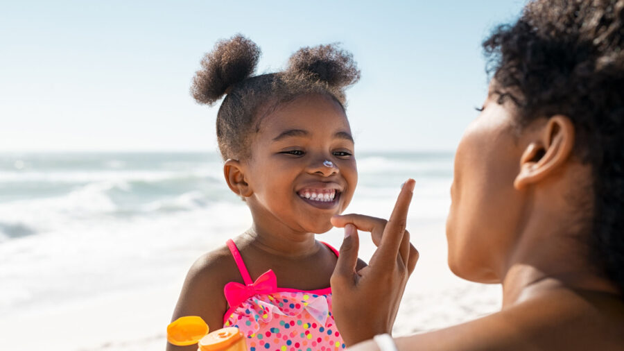 A child in a polka-dot swimsuit smiles at the beach as an adult applies sunscreen to her nose, showcasing one of the important summer safety tips.