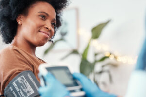 A woman smiles while having her blood pressure checked by a healthcare professional wearing blue gloves, indoors with a blurred plant in the background.