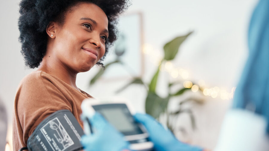 A woman smiles while having her blood pressure checked by a healthcare professional wearing blue gloves, indoors with a blurred plant in the background.