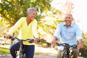 Two older adults ride bicycles outside on a sunny day, smiling and laughing together. Trees with green and autumn-colored leaves are in the background, demonstrating the joy that comes from following healthy aging tips and staying active outdoors.