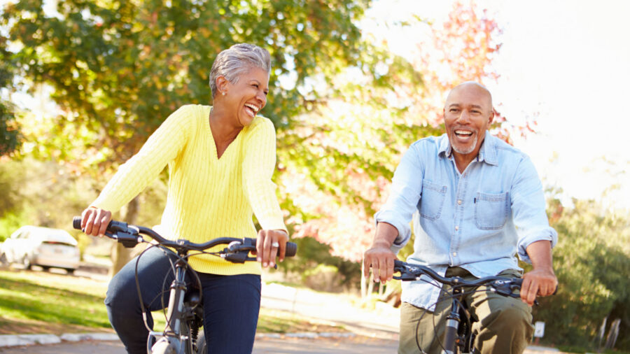 Two older adults ride bicycles outside on a sunny day, smiling and laughing together. Trees with green and autumn-colored leaves are in the background, demonstrating the joy that comes from following healthy aging tips and staying active outdoors.